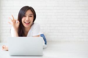 Beautiful portrait asian young woman working online laptop with excited lying on floor brick cement background, freelance girl smile using notebook computer with happy, business and lifestyle concept. photo