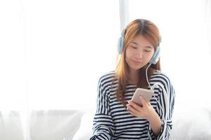 Beautiful asian young woman enjoy listen music with headphone and holding smart mobile phone while sitting in bedroom, relax girl with earphone, leisure and technology concept. photo