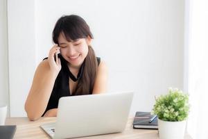 Beautiful young freelance asian woman smiling working on laptop computer and talking mobile smart phone at desk office with professional, girl using notebook, business and lifestyle concept. photo