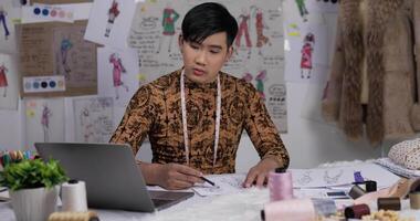 portrait d'un homme de créateur de vêtements asiatique travaillant sur un ordinateur portable et dessinant un croquis de vêtements dans le studio. startup petit entrepreneur est en train de créer une nouvelle collection de vêtements. video