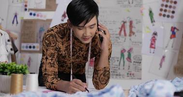Portrait of Asian fashion designer man talking on mobile phone with customer and drawing a sketch clothing at the studio. Startup small business owner. SME marketing and entrepreneur concept.