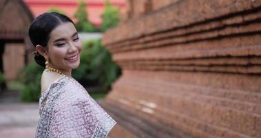vista traseira da mulher tailandesa em traje tradicional, olhando para a câmera e andando no templo antigo.
