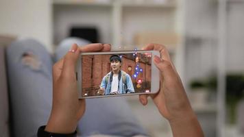 feche o celular da mulher assistindo smartphone em casa. blogueiro de viagens de homem asiático transmitindo vídeo ao vivo na tela do celular com muitos emojis e amor emocional. video