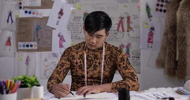 Portrait of Asian Tired fashion designer man draws a sketch clothing while sitting at the studio. Startup small businessman is in process of creating a new clothes collection. video