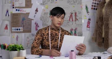 stilista maschio asiatico che controlla i dati su carta o contempla i disegni mentre è seduto sul ponte. stilista di abbigliamento uomo concentrato che lavora nel suo ufficio. video
