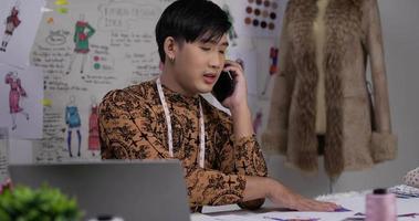 portrait d'un créateur de vêtements asiatique travaillant sur un ordinateur portable et parlant sur mobile avec un client dans le studio. startup petit entrepreneur est en train de créer une nouvelle collection de vêtements. video
