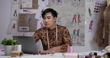 retrato de homem de designer de roupas asiáticas trabalhando no laptop e desenhando uma roupa de esboço no estúdio. startup pequeno empresário está em processo de criação de uma nova coleção de roupas. video