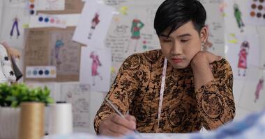 Portrait of Asian Tired fashion designer man draws a sketch clothing while sitting at the studio. Startup small businessman is in process of creating a new clothes collection. video