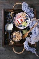 Smoothie bowl healthy breakfast. Strawberry yogurt with blueberries, granola, mint, jug of milk and carambola in wooden tray on textile gauze over dark metal texture background. Top view