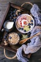 Smoothie bowl healthy breakfast. Strawberry yogurt with blueberries, granola, mint, jug of milk and carambola in wooden tray on textile gauze over dark metal texture background. Top view