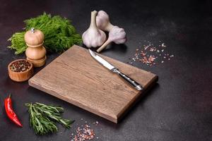 A wooden cutting board with spices, herbs, cherry tomatoes and salt on a black concrete background