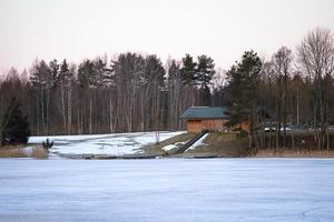 A scenic view of a bathhouse on a bank of a white frozen lake with stairs to the lake and trees round it photo