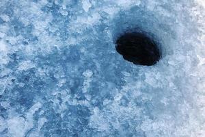 agujero de hielo de agua redondo oscuro para la pesca en hielo hecho en hielo grueso de lago con nieve azul dura en la superficie foto
