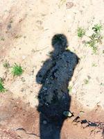 Long shadow of a person in jacket with hood on dry sand with growing green grass and lots of stones and rocks photo