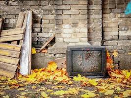 Old broken TV set covered in dirt with small red heart standing outdoors in yellow autumn maple leaves near brick wall and wooden planks an other trash photo