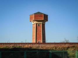 Red brick old water tower on blue sky over railway building behind an iron fence photo