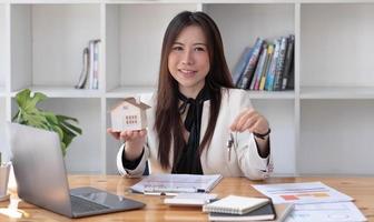 casa en miniatura en manos de una mujer asiática agente de bienes raíces préstamo hipotecario que trabaja en la oficina. mirando a la cámara. foto