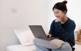 retrato de una joven asiática descansando con una laptop en casa, sentada en un sofá en una sala de estar moderna. foto