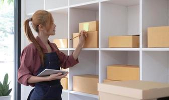 Young business woman working online e-commerce shopping at her shop. Young woman seller prepare parcel box of product for deliver to customer. Online selling, sme, e-commerce. photo