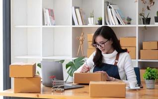 Young beautiful happy asian business woman owner of SME online using laptop receive order from customer with parcel box packaging at her startup home office, online business seller and delivery photo