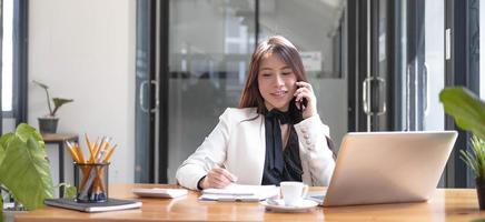 hermosa mujer asiática joven sentada en la cafetería usando un teléfono inteligente. feliz joven mujer de negocios sentada a la mesa en un café con una computadora con pestaña. foto