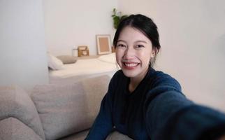 Portrait of Young Asian woman video call with her friend and checking social media by smartphone sitting on couch. Lifestlye concept. photo