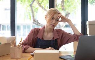 A portrait of young serious Asian woman working with laptop in the office full of packages and boxes stacking up, busy looking table, for SME, delivery, start up business and home office concept. photo