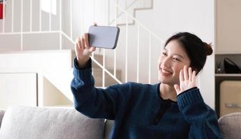 una joven asiática sonriente usando un teléfono móvil mientras se sienta en un sofá en casa con una computadora portátil foto