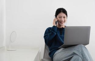 portrait of a happy casual asian woman holding laptop computer and hold smartphone  while sitting on a chair over white background photo