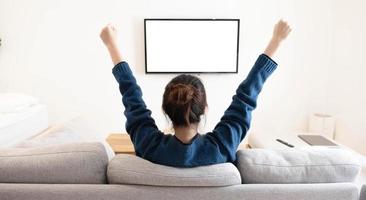detrás de una joven asiática viendo televisión de pantalla blanca con victoria en el sofá en la sala de estar en casa foto