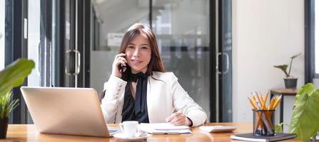 encantadora mujer asiática que trabaja en la oficina usando un teléfono inteligente y una computadora portátil mirando la cámara. foto