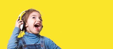 Little girl in a striped blue turtleneck listens to music with white headphones and sings loudly with her mouth open. Fun, shouting, children's emotions. Yellow background, banner, copy space