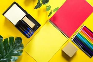 Layout of multi-colored stationery on a yellow background spiral notebook, colored pencils, stand, pens, indoor flower. Business flat lay, mock up. Sheets for notes, office work. Back to school photo