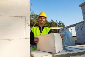 el constructor tiene en sus manos un bloque de hormigón celular: la mampostería de las paredes de la casa. trabajadores de la construcción con ropa protectora: un casco y un chaleco reflectante. foto