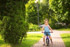 una chica con un traje de mezclilla monta una bicicleta morada en verano en un parque verde. entretenimiento activo para niños, scooter para niños pequeños, bebé feliz. copie el espacio foto