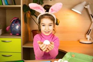 A girl in the ears of an Easter bunny paints eggs with a felt-tip pen in the home interior. Crafts, preparation for a religious holiday, a tray with eggs, hare ears made of plasticine photo