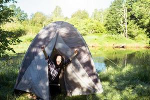 A happy woman in a plaid shirt looks out of a tourist tent on a hike on the riverbank in the morning. Camping in nature, overnight in the wild, family holidays and adventures. photo