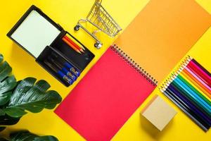 Layout of multi-colored stationery on a yellow background spiral notebook, colored pencils, stand, pens, indoor flower. Business flat lay, mock up. Sheets for notes, office work. Back to school photo