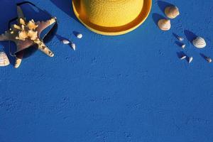 marco con accesorios de playa sobre un tema náutico sombrero de paja amarillo, gafas de sol, estrellas de mar y conchas sobre un fondo azul. concepto de vacaciones, viaje por mar, protección uv, natación. copie el espacio plano foto