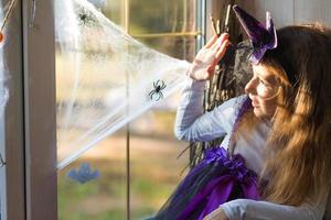 A girl in a witch costume on the windowsill by the window is playing with a spider web and a spider, decorating the house for the Halloween holiday. Terrible scenery, fear and horror, pumpkins photo