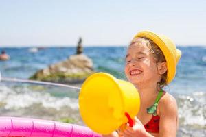 chica con sombrero de paja amarillo juega con el viento, el agua y un dispensador de agua en una piscina inflable en la playa. productos indelebles para proteger la piel de los niños del sol, las quemaduras solares. recurso en el mar. foto