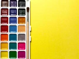 A palette of eighteen-color watercolors and paintbrushes on a yellow background with space for text. photo