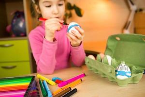 Girl with bunny ears makes medical masks out of plasticine for Easter  eggs. An egg with eyes and a blue mask in the tray. DIY sitting at home, preparing for a religious holiday, home interior. photo