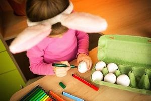 A girl in the ears of an Easter bunny paints eggs with a felt-tip pen in the home interior. Crafts, preparation for a religious holiday, a tray with eggs, hare ears made of plasticine photo