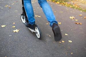 The legs of a man in jeans and sneakers on a scooter in the park in autumn with fallen dry yellow leaves on the asphalt. Autumn walks, active lifestyle, eco-friendly transport, traffic photo