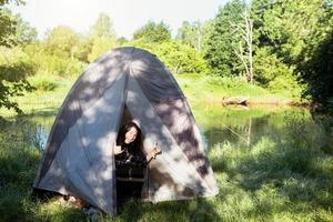 A happy woman in a plaid shirt looks out of a tourist tent on a hike on the riverbank in the morning. Camping in nature, overnight in the wild, family holidays and adventures. photo