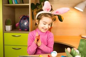 A girl in the ears of an Easter bunny paints eggs with a felt-tip pen in the home interior. Crafts, preparation for a religious holiday, a tray with eggs, hare ears made of plasticine photo