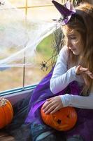 una niña disfrazada de bruja en el alféizar de la ventana está jugando con una telaraña y una araña, decorando la casa para la fiesta de halloween. terrible paisaje, miedo y horror, calabazas