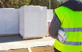 The project architect's back is on the construction site of a house with the foundation laid and the blocks delivered. Construction worker in a protective yellow helmet and a signal vest. Mock up photo