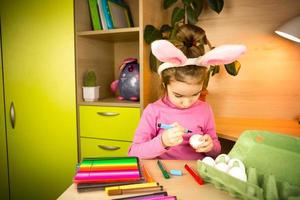 A girl in the ears of an Easter bunny paints eggs with a felt-tip pen in the home interior. Crafts, preparation for a religious holiday, a tray with eggs, hare ears made of plasticine photo